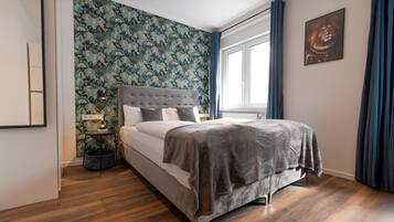 A bedroom with a bed, a nightstand, a wall with a floral wallpaper, a window with curtains, and a painting on the wall.