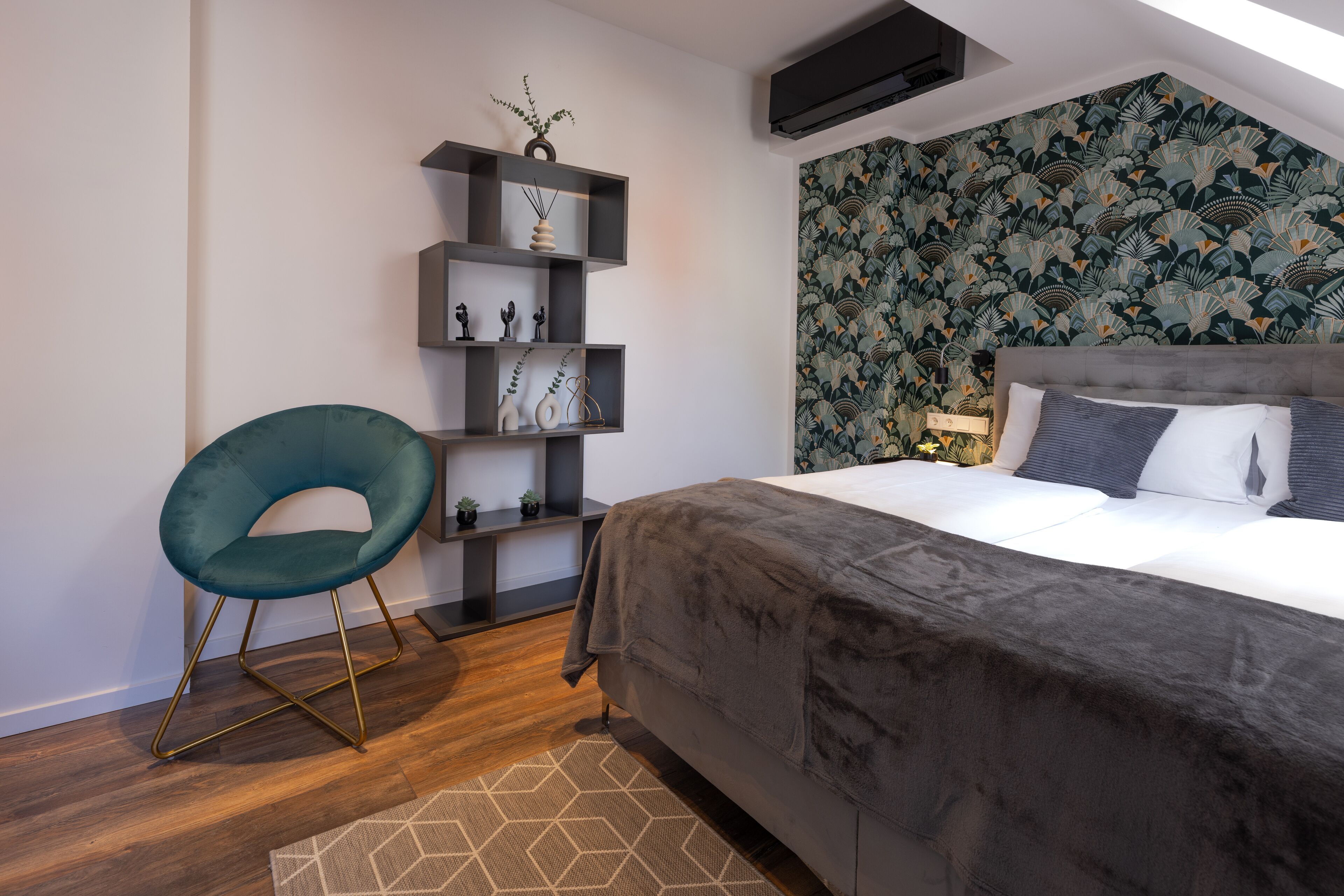 A modern bedroom with a grey upholstered bed, a teal chair, and a wall-mounted shelf with decorative items.