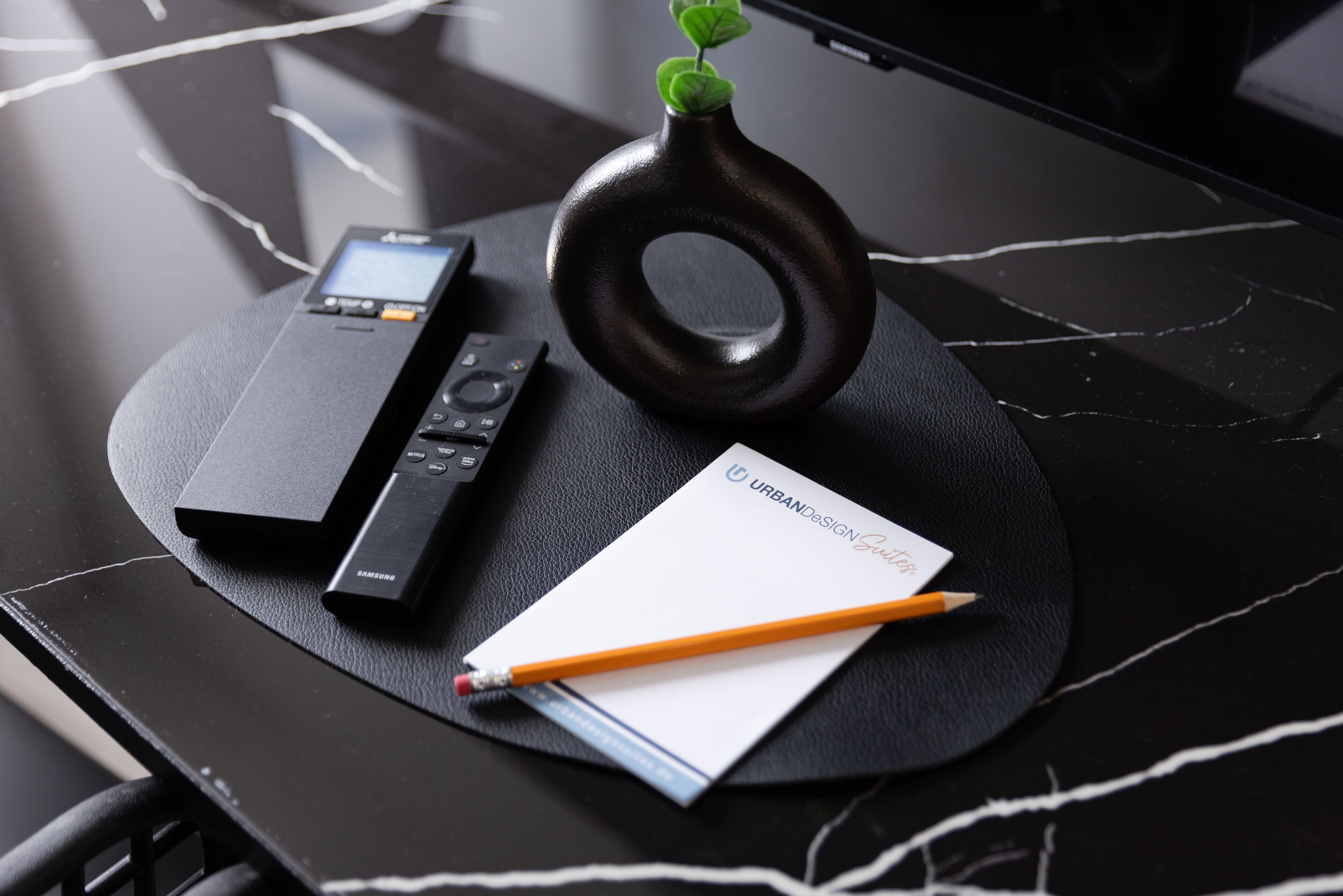 Ein schwarzer Tisch mit einem Samsung-Gerät, einem Notizblock, einem Bleistift und einer dekorativen Vase mit Pflanze.