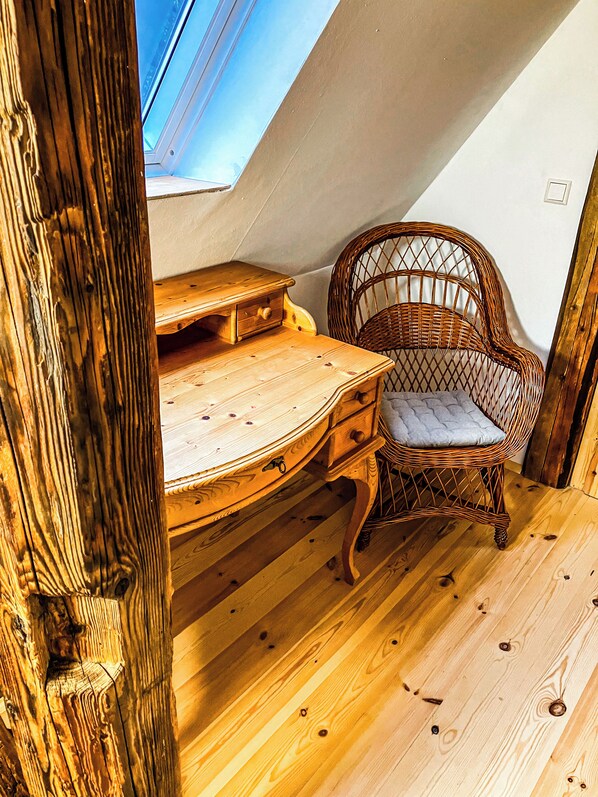 Une pièce avec un plancher en bois et un plafond en pente, où l’on trouve un bureau en bois et une chaise en osier.