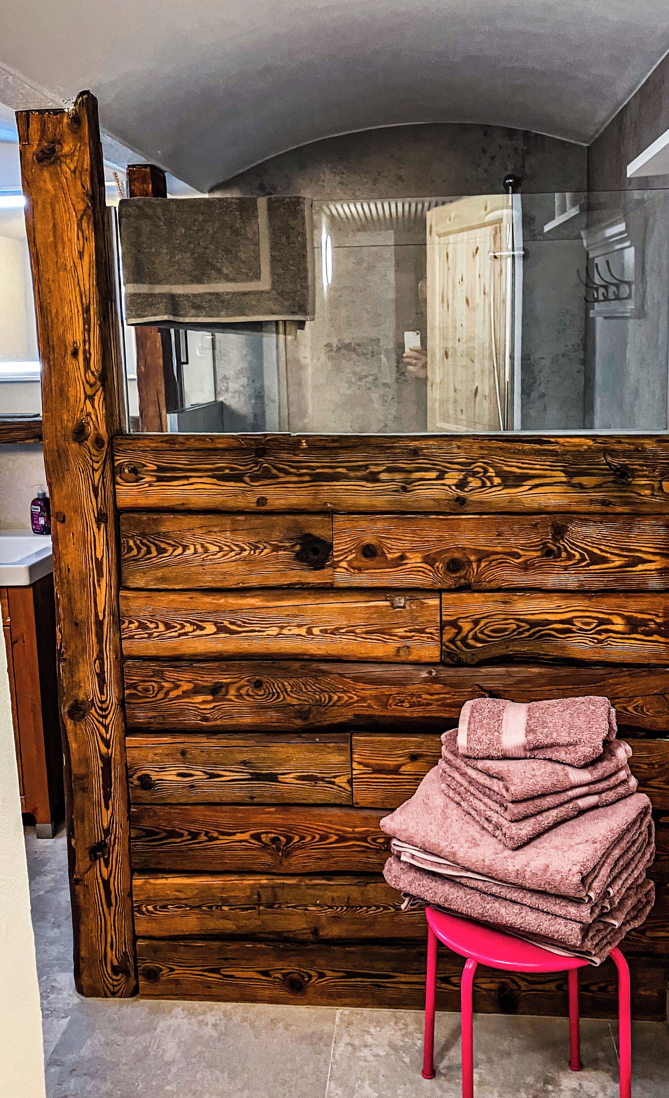 A bathroom with a wooden partition, a stack of towels on a red stool, and a mirror reflecting a shower area.
