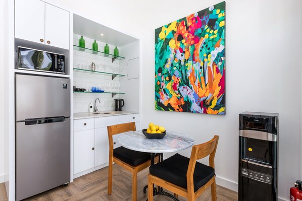 A modern kitchen with a stainless steel refrigerator, a microwave, and a water dispenser. There is a dining table with two chairs and a bowl of fruit. A colorful abstract painting is on the wall.