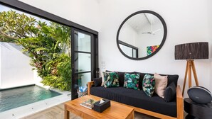 A modern living room with a black sofa, a wooden coffee table, a round mirror, and a floor lamp. There is a small pool visible through a glass door. - Villa Jabula  (Seminyak)
