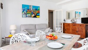 A dining area with a table set for two, a grey sofa, and a colorful cityscape painting on the wall. - Atic Cap Tossa by Interhome (Tossa de Mar)