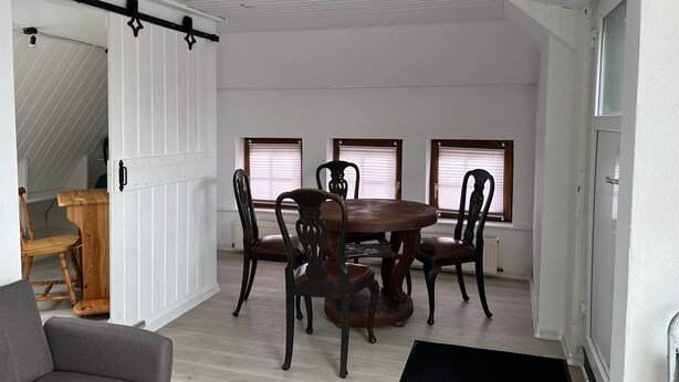 Et spiseområde med et rundt bord, seks stole, et træsidebord, en grå sofa og en sort måtte.