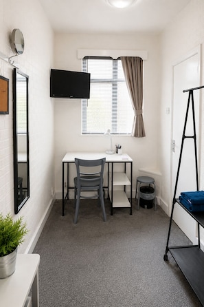Una habitación pequeña y limpia con escritorio, silla, televisión, ventana, cortinas y una planta pequeña.
