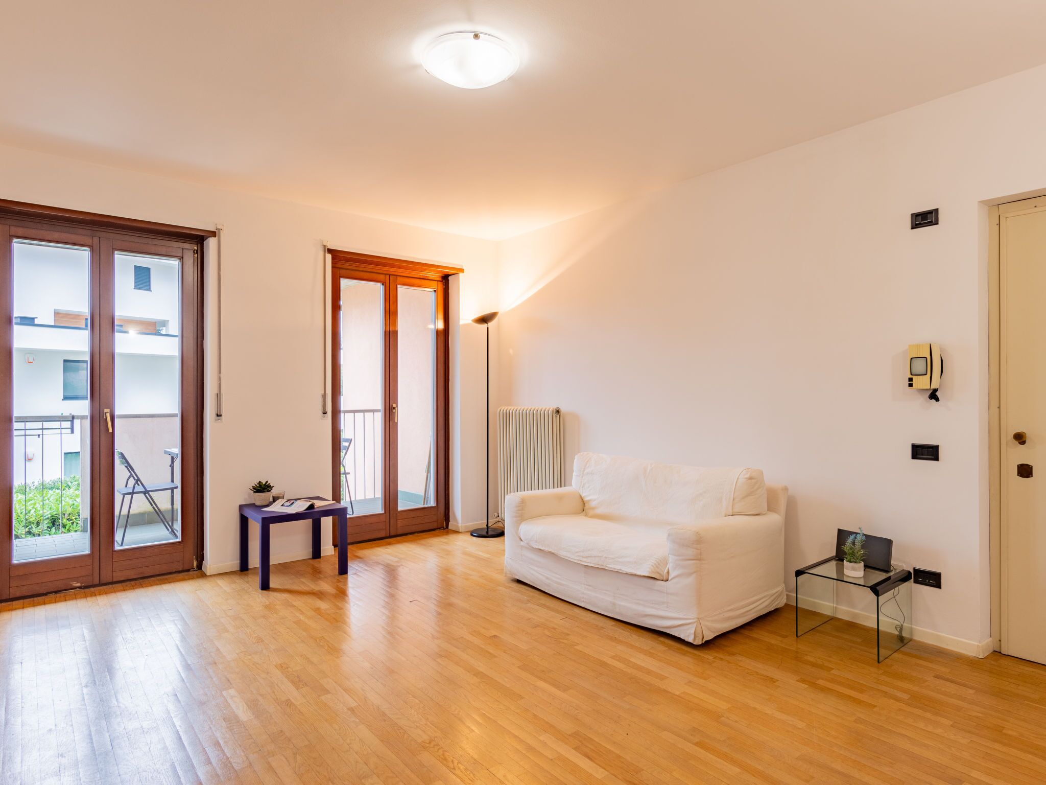 A spacious living room with a wooden floor, a white sofa, a glass coffee table, and a sliding glass door leading to a balcony.