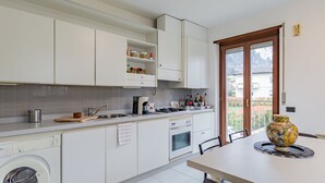 Une cuisine moderne avec des meubles blancs, un lave-linge encastré, un coin repas avec une table et des chaises, et une vue sur l’extérieur grâce à une porte vitrée.