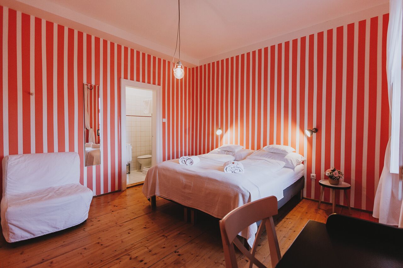 A hotel room with striped wallpaper, a bed with towels, a chair, a small table with a plant, and a bathroom visible through an open door.