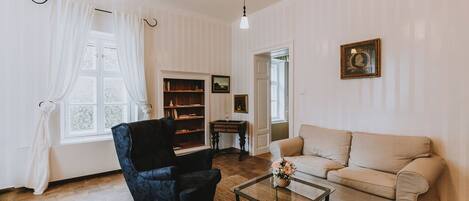 A living room with a dark blue armchair, a beige sofa, a glass coffee table, a bookshelf, and a framed picture on the wall.