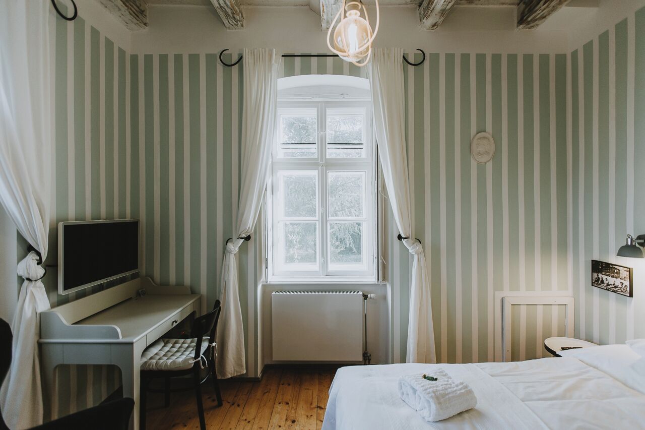 Quarto com cama, escrivaninha e cadeira, janela com cortinas, radiador e luminária fixada na parede.