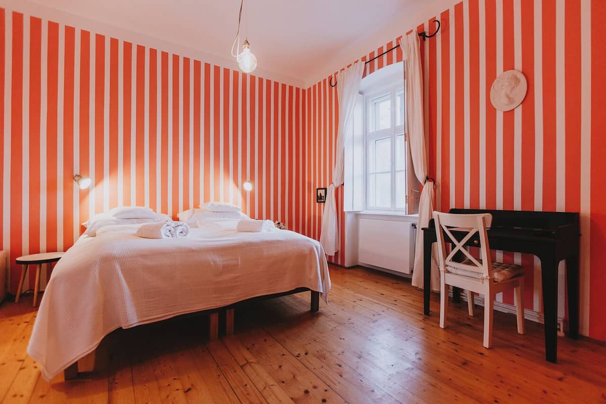 Una habitación con paredes a rayas, una cama con sábanas blancas, un piano y una ventana con cortinas.