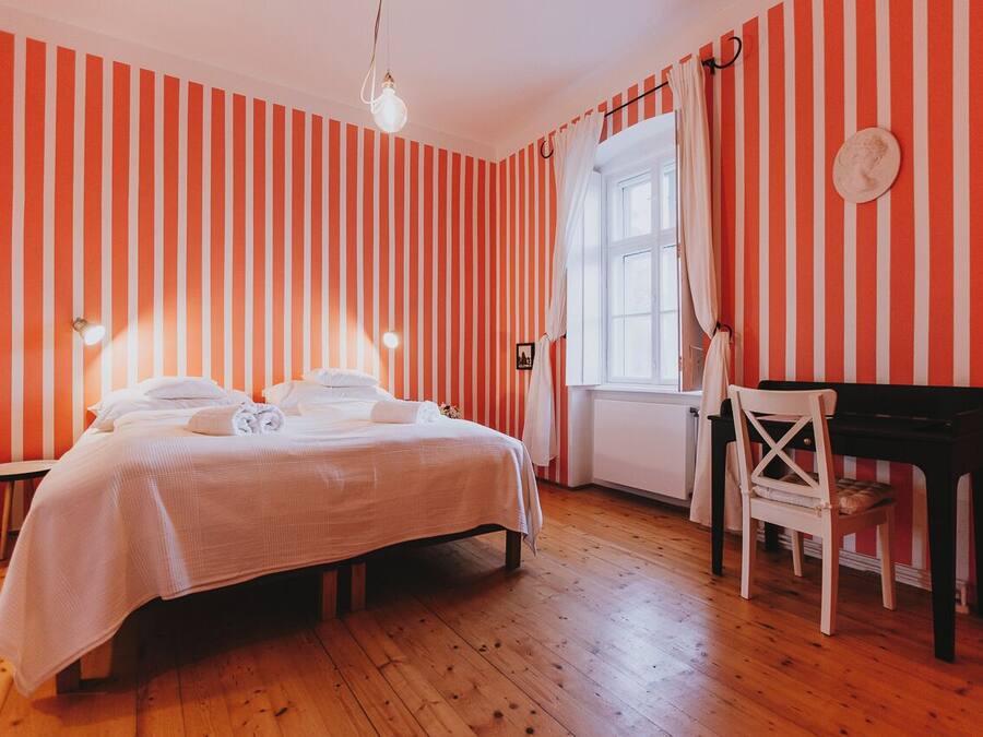 Una habitación con paredes a rayas, una cama con sábanas blancas, un piano y una ventana con cortinas.