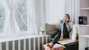 A person sitting on a bench by a window with white curtains, a vase with flowers, and a bookshelf with a book titled "SOPRON". - Villa Mimi (Sopron)