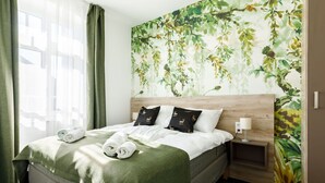Une chambre d’hôtel avec un lit, des rideaux verts, une fresque représentant des feuilles sur un mur, et une table de chevet avec une lampe.