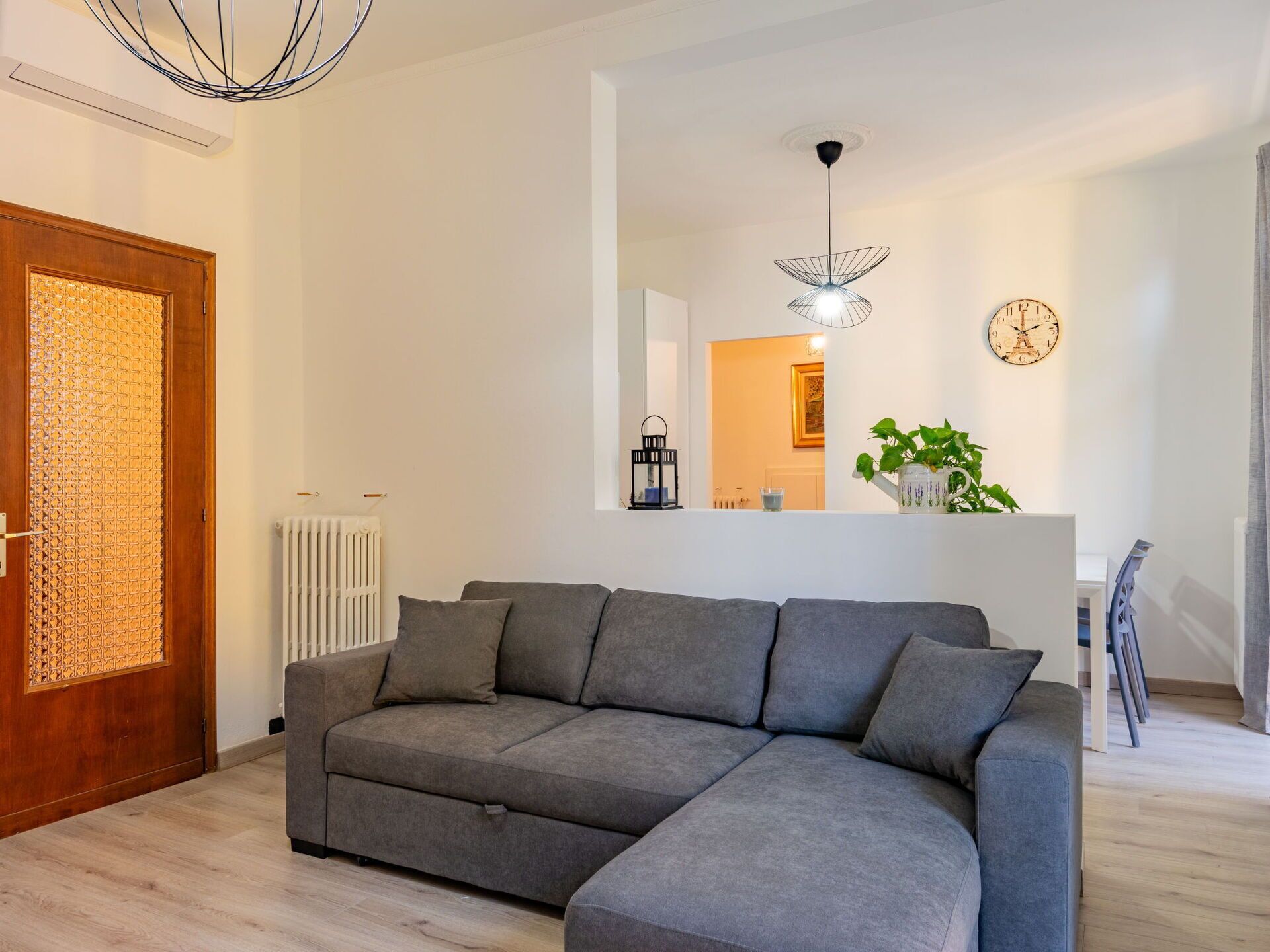 A modern living room with a grey sectional sofa, a wooden door with a decorative panel, a white radiator, a hanging light fixture, a clock, and a plant on a white console table.