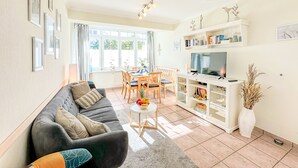 A living room with a grey sofa, a dining table with chairs, a TV on a white stand, and a bookshelf. - nur 100 m bis zum Strand, Zentrumsnahe Ferienwohnung - Villa Meernixe Fewo 10 (Ostseebad Binz)