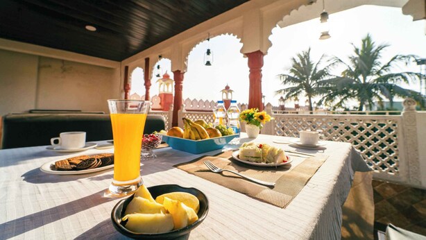 Una mesa de desayuno con un vaso de jugo de naranja, un cuenco de fruta y un plato de comida, en una habitación con vista a palmeras y un arco decorativo.