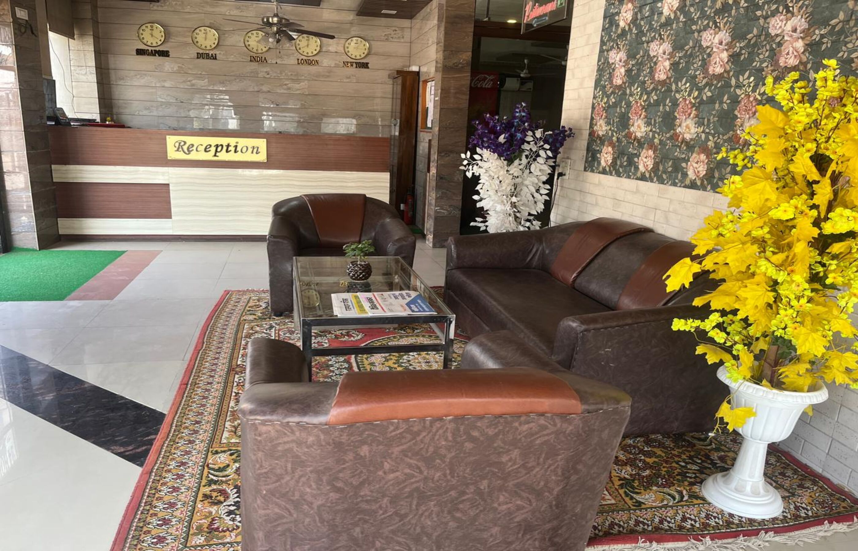 Un área de recepción con mostrador, dos sofás de cuero, una mesa de centro y un jarrón con flores amarillas.