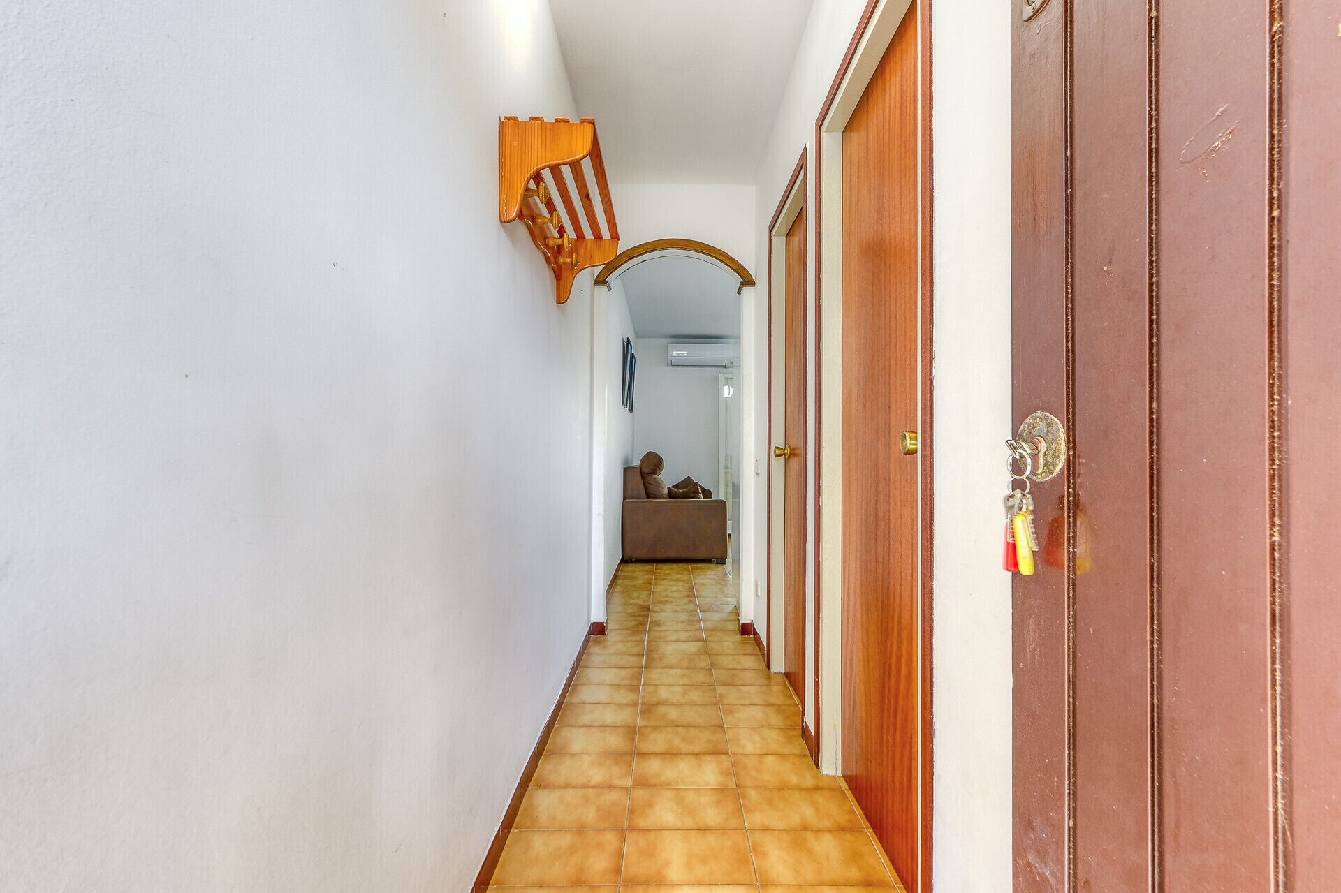 Un couloir avec un sol carrelé, une porte en bois avec un porte-clés, et un canapé marron en arrière-plan.