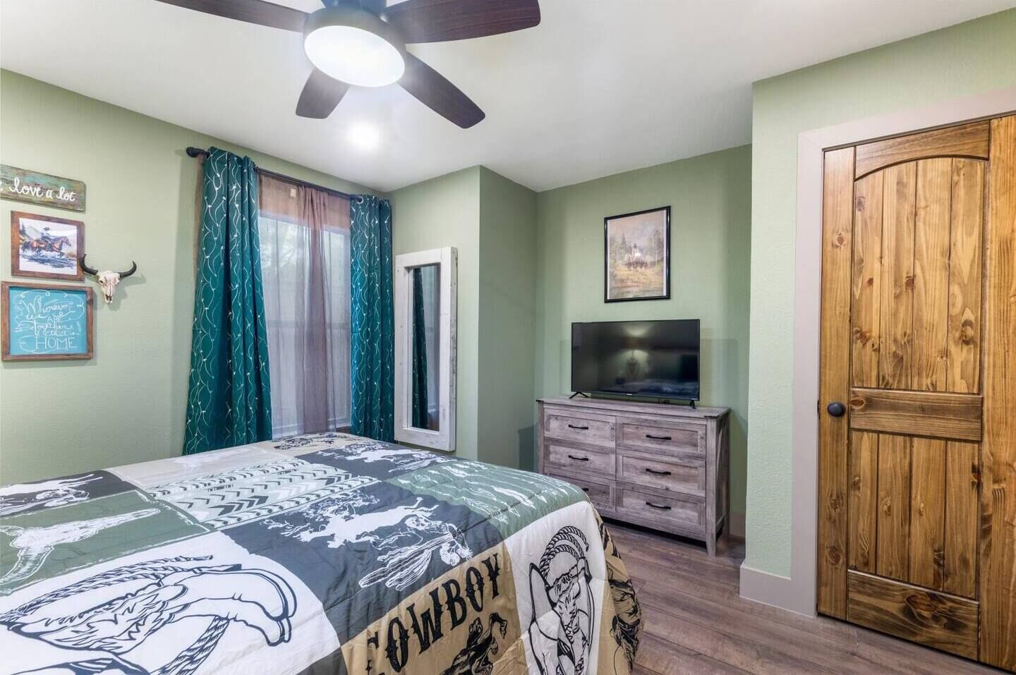 Une chambre à coucher avec un lit, un téléviseur posé sur une commode, une porte en bois et un ventilateur de plafond.