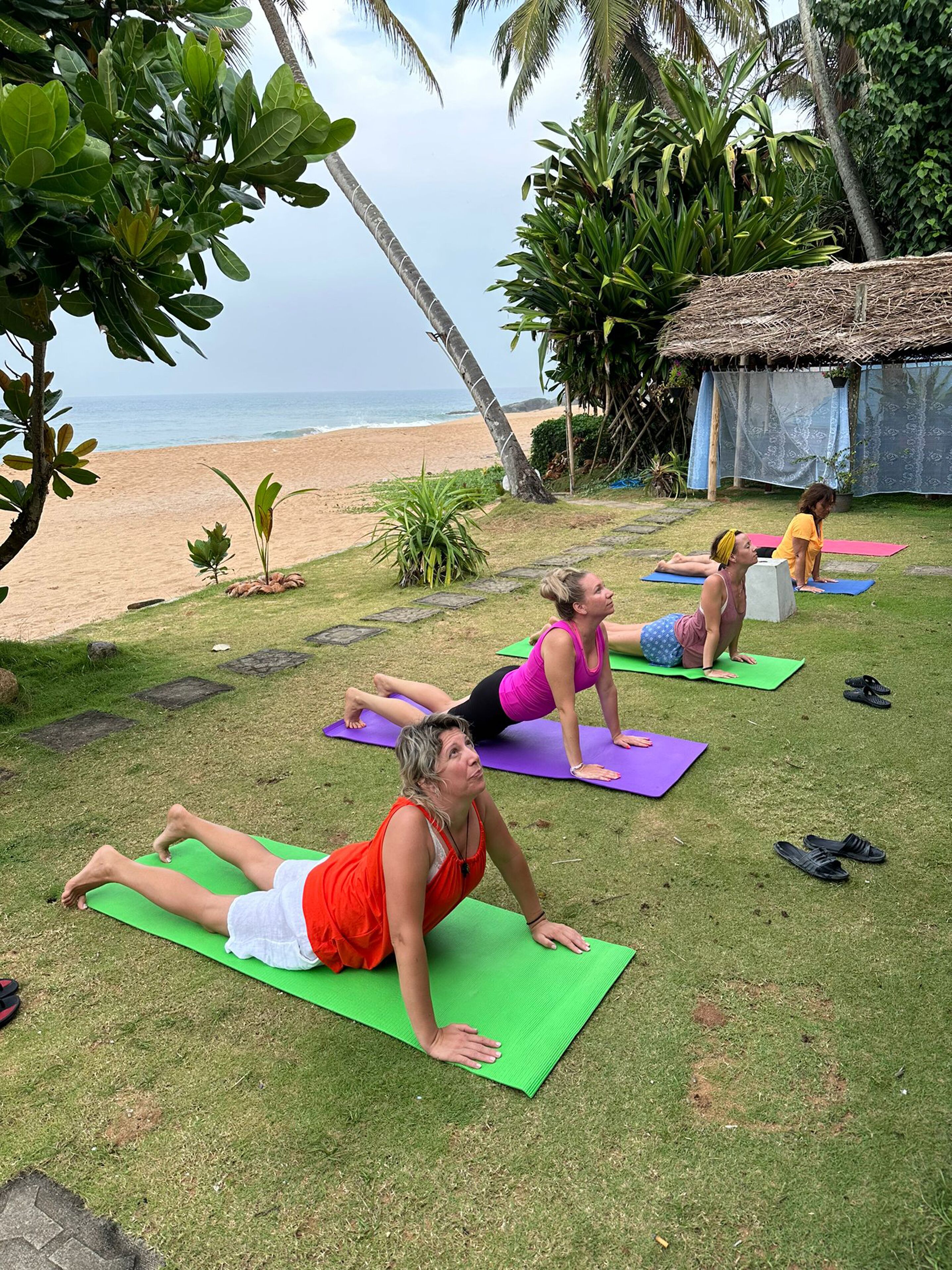 Un grupo de personas practicando yoga sobre esterillas verdes en una playa, con palmeras y una cabaña al fondo.
