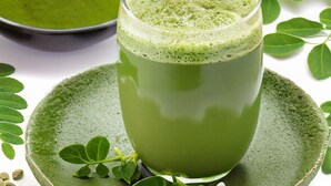 A glass of green beverage on a ceramic plate with a green background, surrounded by green leaves and a bowl of green powder.