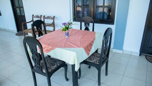 A dining area with a table covered by a pink tablecloth, surrounded by four chairs with black frames and patterned cushions. A vase with flowers is placed on the table.