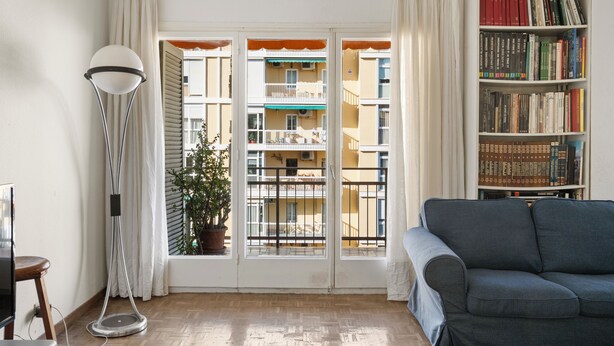 A room with a sofa, a floor lamp, a bookshelf, and a view of balconies through a window.