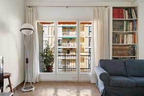A room with a sofa, a floor lamp, a bookshelf, and a view of balconies through a window.