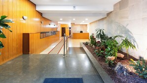 A hotel lobby with a wooden reception desk, a reception area, a staircase, and a plant-lined pathway.