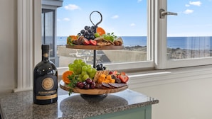 A two-tiered fruit and cheese board with a bottle of wine on a kitchen counter by a window overlooking the sea. - Oceanfront home w/Amazing Views, Hottub! Sleeps 10 (Scituate)