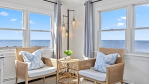 A room with two wicker sofas facing a large window overlooking the sea, a small round table with a plant, and two wall-mounted light fixtures. - Oceanfront home w/Amazing Views, Hottub! Sleeps 10 (Scituate)