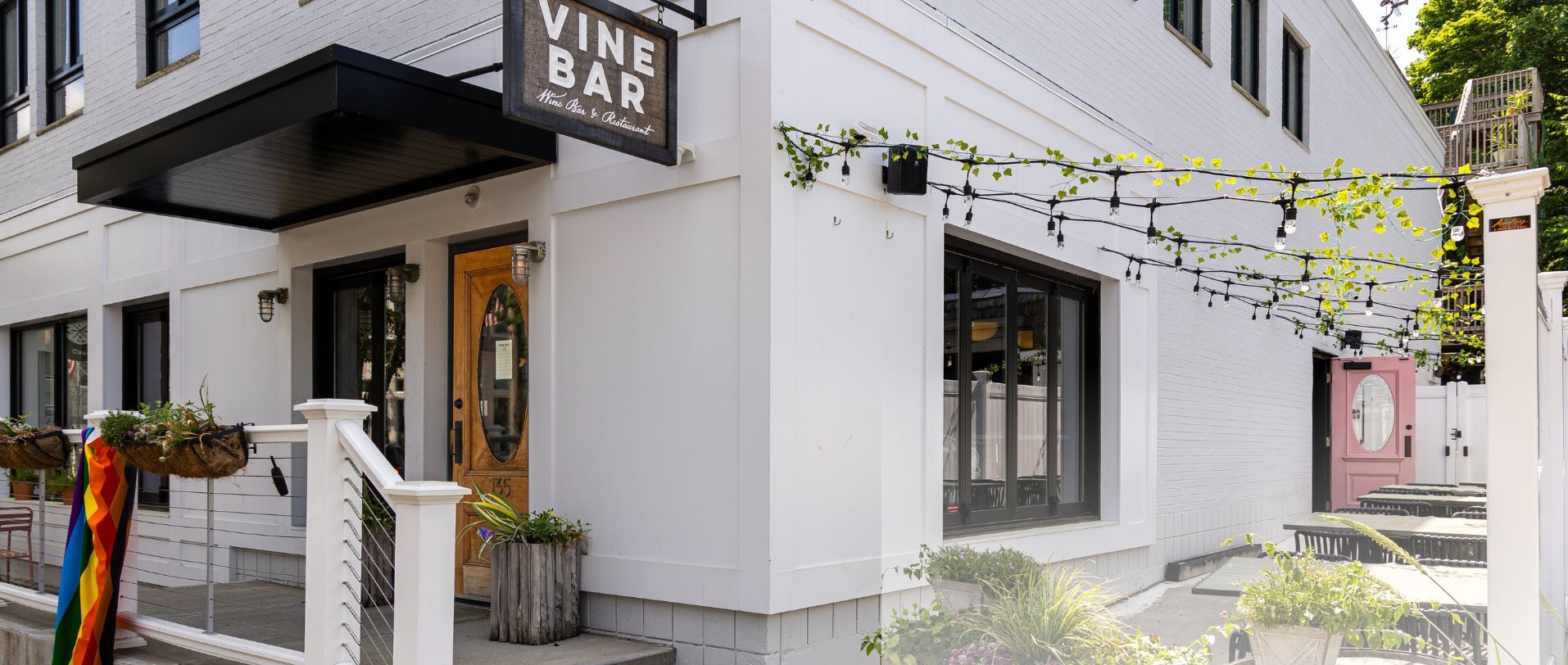 A white building with a 'Vine Bar' sign, outdoor seating, and potted plants.