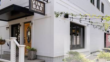 A white building with a 'Vine Bar' sign, outdoor seating, and potted plants.