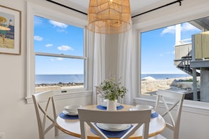 A dining area with a table set for two, a vase with flowers, and a view of the beach and ocean through large windows.