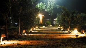 Un sendero nocturno iluminado por lámparas, rodeado de árboles, que lleva a un edificio con techo de paja.