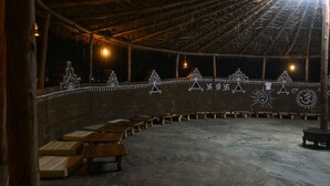 Una estructura al aire libre con techo de paja, bancos de madera y paredes decoradas con diseños geométricos y simbólicos.