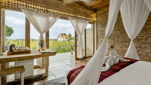 A room with a bed, a table, and a view of a house and field.