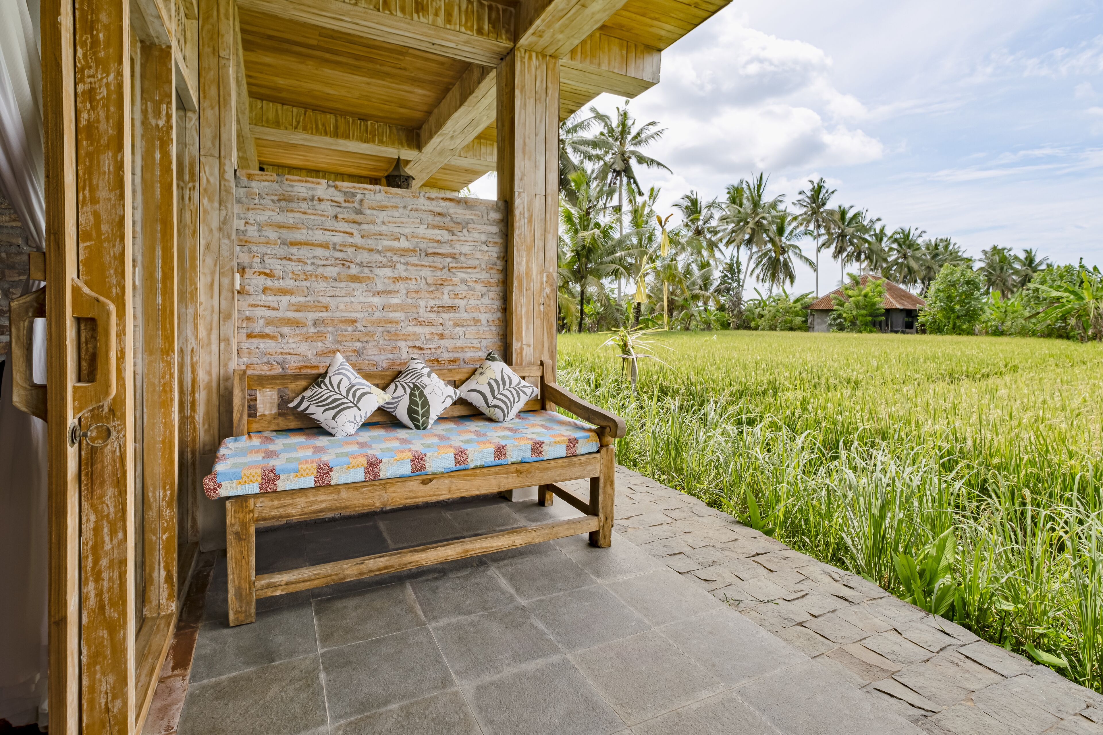 A wooden bench with patterned cushions under a wooden structure with a brick wall, adjacent to a lush green field and palm trees.