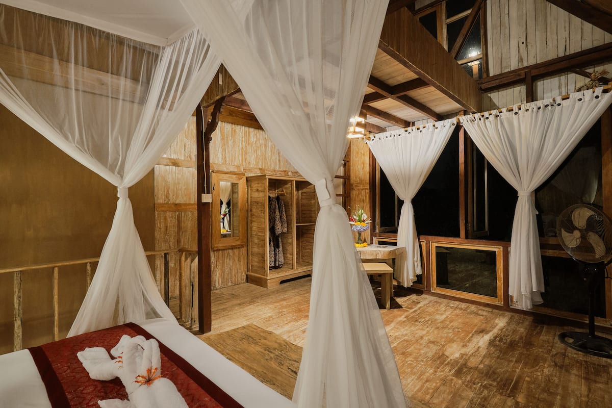 Una habitación con cama, dosel blanco, paredes de madera, puerta de madera, ventilador y ventana.
