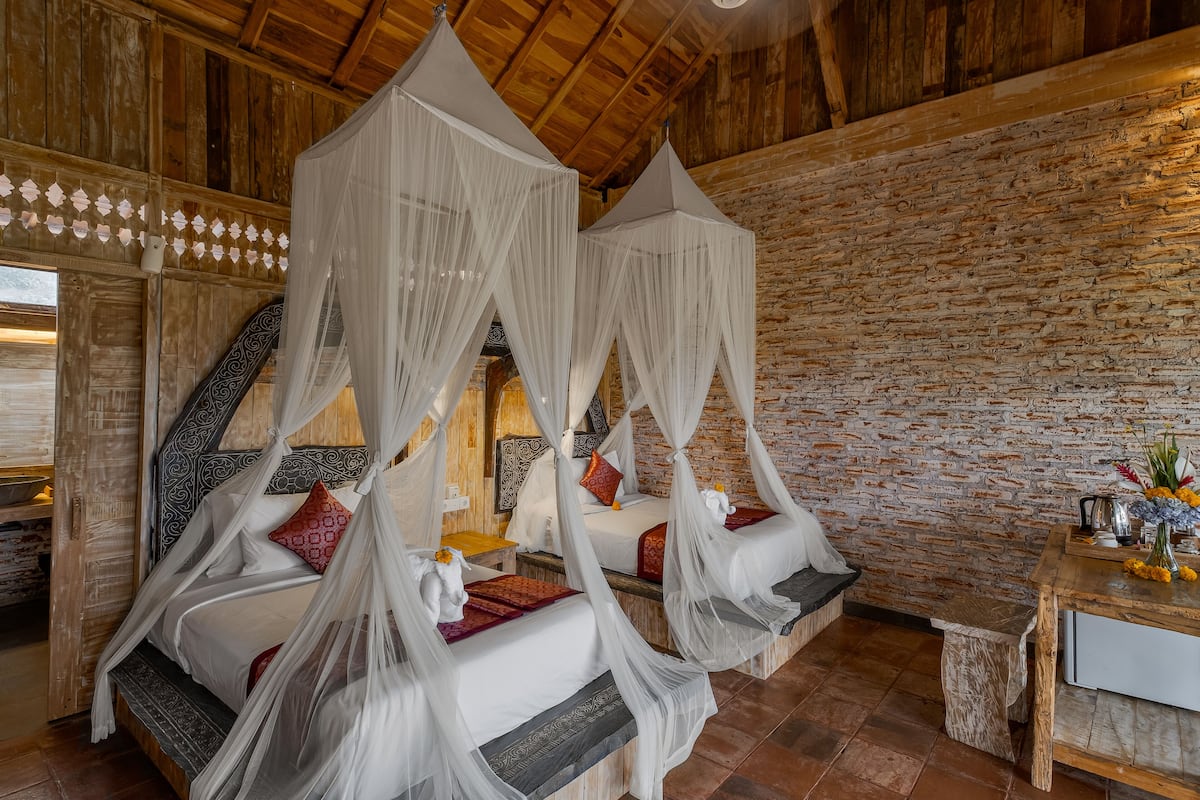 Una habitación con dos camas, cada una con dosel. La habitación tiene un techo de madera rústica y paredes de piedra. A la derecha, hay una pequeña mesa con un jarrón de flores.