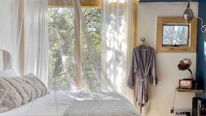 A bedroom with a large bed, a window with sheer curtains, a bedside lamp, and a small table with a radio.