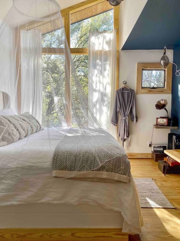 Une chambre avec un grand lit, une fenêtre avec des rideaux légers, une lampe de chevet et une petite table sur laquelle se trouve une radio.