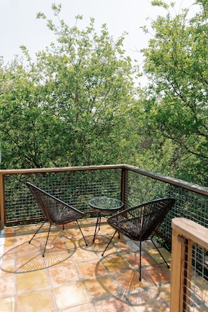 Un balcon avec deux chaises et une table, entouré d’une rambarde en métal, avec des arbres en arrière-plan.