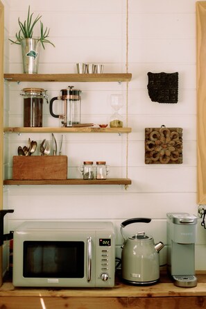 A kitchen with a microwave, kettle, and coffee maker on the counter. There are shelves with utensils, jars, and a plant.