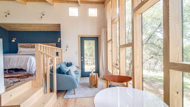 A cozy room with a bed, a blue sofa, a small table, and large windows offering a view of the outdoors. - Sapling Treehouse at HoneyTree (Fredericksburg)