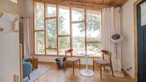 A room with large windows, wooden flooring, a table, two chairs, a fan, and a basket with items. - Sapling Treehouse at HoneyTree (Fredericksburg)