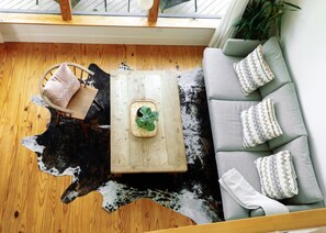 A room with a wooden floor, a sofa with patterned cushions, a small wooden table with a plant, and a chair with a cushion. - Sycamore Treehouse at The Meadow by HoneyTree (Fredericksburg)