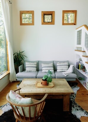 A cozy living room with a grey sofa, a wooden coffee table, and a potted plant. There are three framed pictures on the wall, and a staircase is visible to the right. - Sycamore Treehouse at The Meadow by HoneyTree (Fredericksburg)